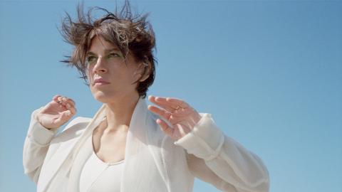 Woman in a white suit, a blue sky her background. She is visible only from the chest up.  She is looking off camera, her hands raised up to her shoulders, elbows back, palms facing out.  Her short brown hair is blowing up in the wind.
