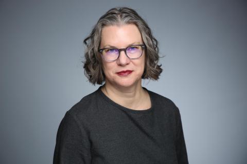 Portrait of a white woman, seated and turned 3/4 towards the camera. She is wearing dark grey glasses and a black top.
