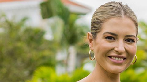 a woman, seen from the neck up, smiling at the camera, standing with lush greenery in the background.