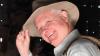 A close up image of a man’s face in three-quarter profile, smiling as he touches the brim of his hat. He seems to be dressed for travel and adventure.